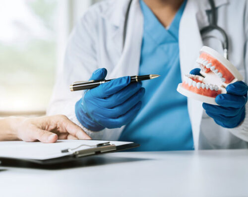 A dentist wearing blue gloves explains a dental procedure using a tooth model, while a patient takes notes on a clipboard, illustrating a consultation session.