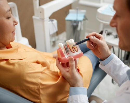 Closeup of male dentist holding tooth model while explaining dental implant surgery to a new patient in a dental clinic.