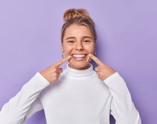 Happy young woman pointing at her teeth and smiling, showcasing a healthy and bright smile against a purple background.